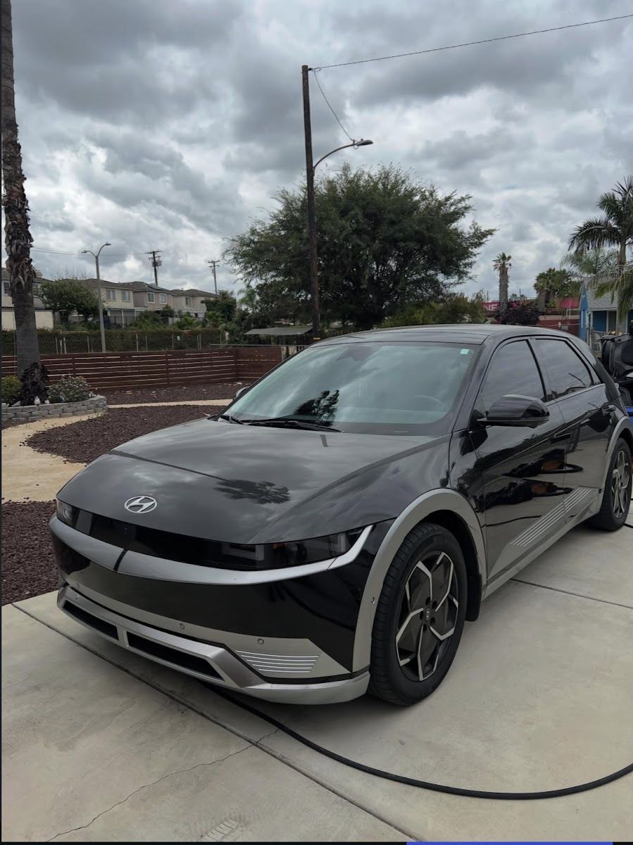 A black Hyundai Ioniq 5 being washed on a driveway, hose laid out, under an overcast sky